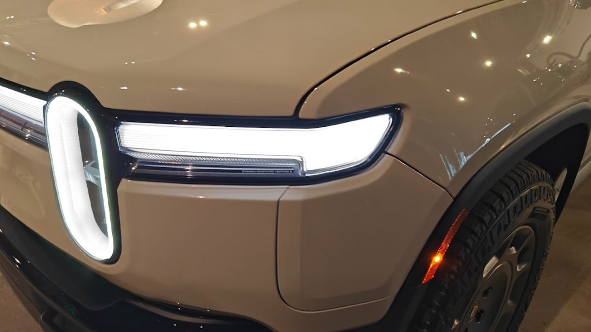 Rivian R1T electric SUV in cream white, front quarter view, showcasing distinctive Thor's Hammer LED headlights inside showroom with charging information display.