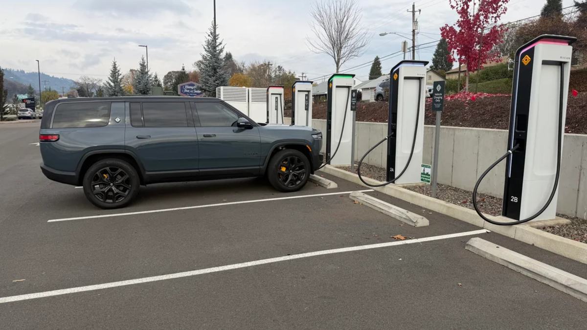A gray 2024 Rivian R1S is shown from the side view at an EV charging station, featuring black wheels and positioned next to multiple charging pedestals.