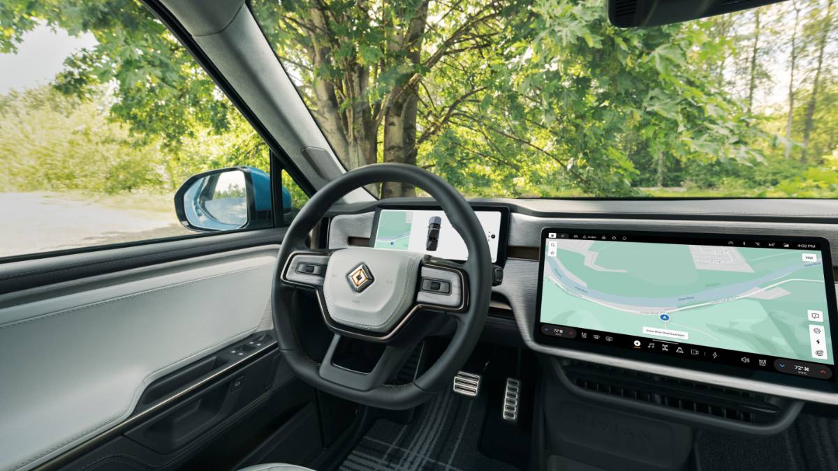 Car interior showing steering wheel and two digital screens displaying navigation maps. Trees outside.