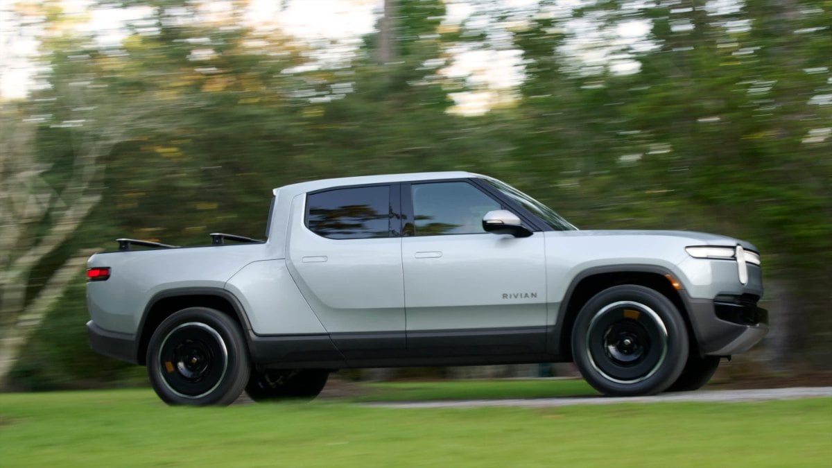 White 2025 Rivian R1T electric pickup truck driving on a tree-lined road with motion blur, side profile view
