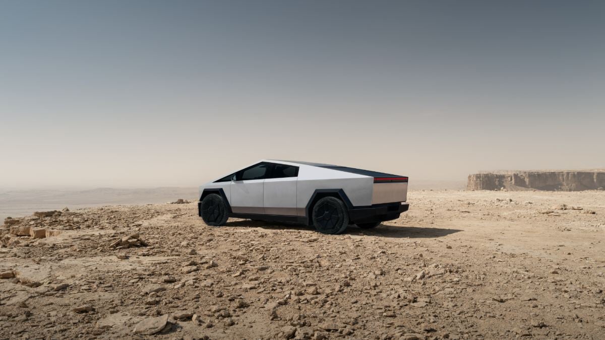 A silver Tesla Cybertruck shown from a side 3/4 angle, positioned in a stark desert landscape with rocky terrain and cliffs in the background.
