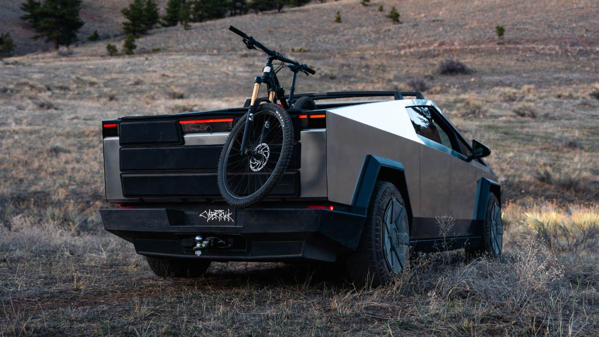 A Tesla Cybertruck shown from the rear quarter view in matte black/silver, with a mountain bike mounted on top, photographed in a rugged outdoor setting.