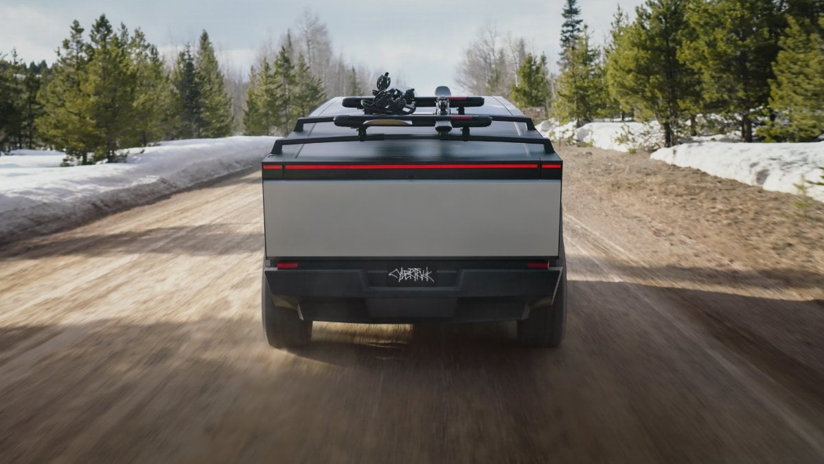 Rear view of a silver Tesla Cybertruck driving on a snowy forest road, equipped with roof-mounted ski/snowboard racks and showing its distinctive angular rear design and full-width LED light bar.