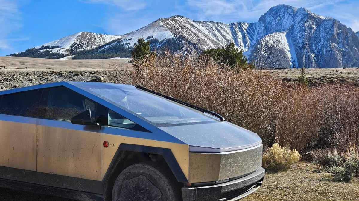 A stainless steel Tesla Cybertruck photographed from the front quarter angle against a dramatic backdrop of snow-capped mountains, showcasing its distinctive angular design and futuristic appearance.