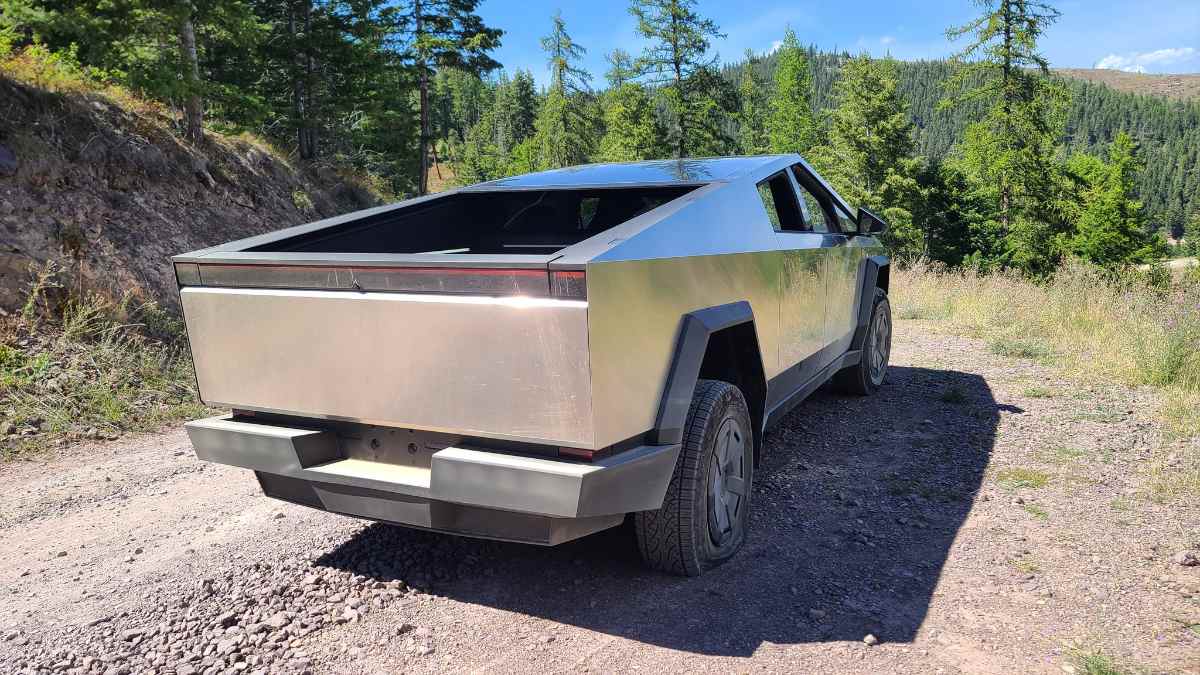 Stainless steel 2025 Tesla Cybertruck parked on gravel road with pine forest backdrop, angular geometric design gleaming in sunlight