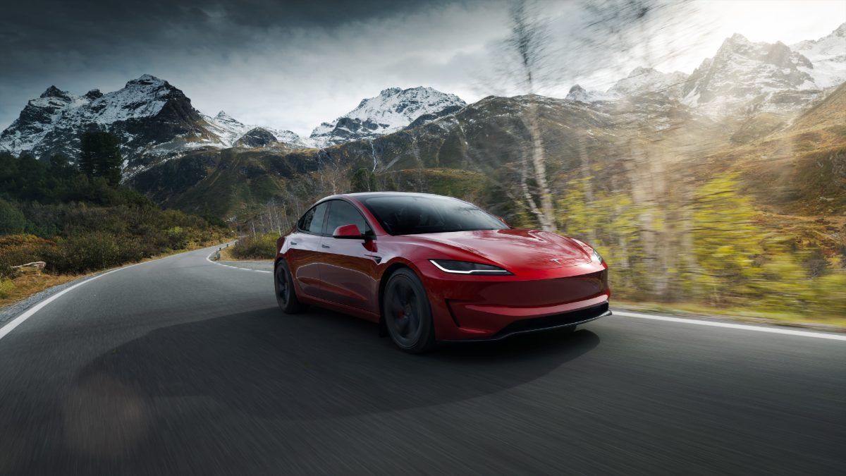 Candy red Tesla Model 3 driving along winding mountain road with snow-capped peaks and autumn trees