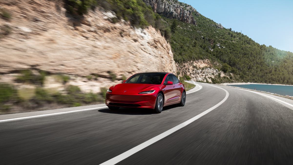 Candy red Tesla Model 3 cruising on a winding mountain road with rocky cliffs and forest backdrop