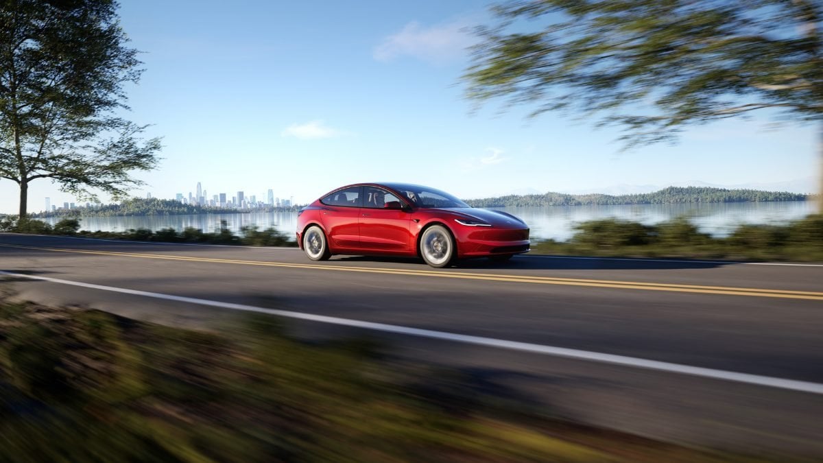 Red car driving on a lakeside road with a city skyline in the background.