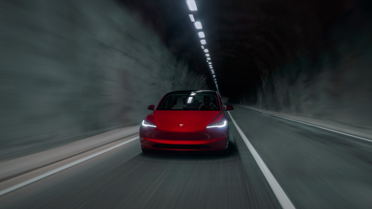 Bright red Tesla driving through dimly lit tunnel with headlights illuminating gray concrete walls
