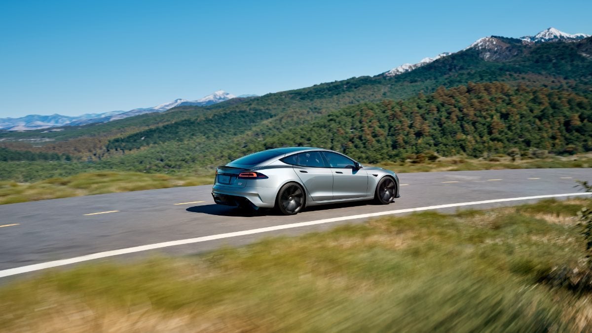Silver car driving on a scenic mountain road with green hills and blue sky.