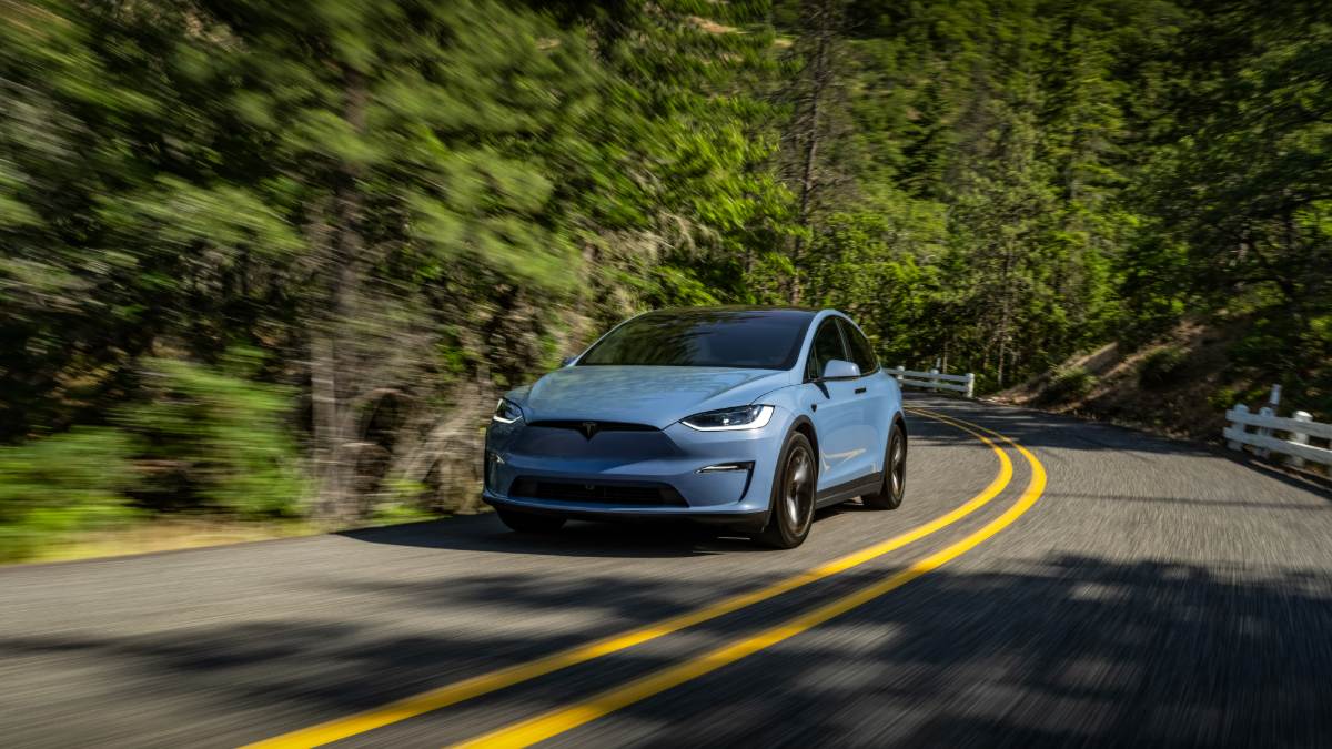Light blue Tesla Model X SUV, front three-quarter view, driving on curved forest road with motion blur and yellow lines.