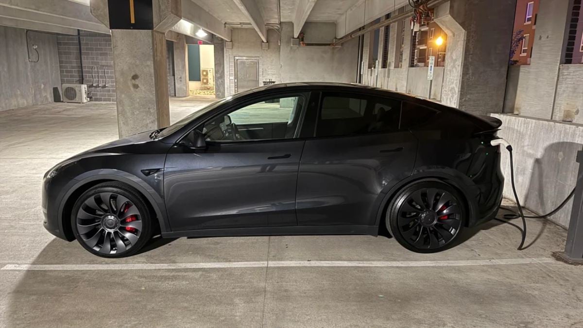 Black 2025 Tesla Model Y charging in minimalist concrete parking garage, side profile with defined lines and black wheels