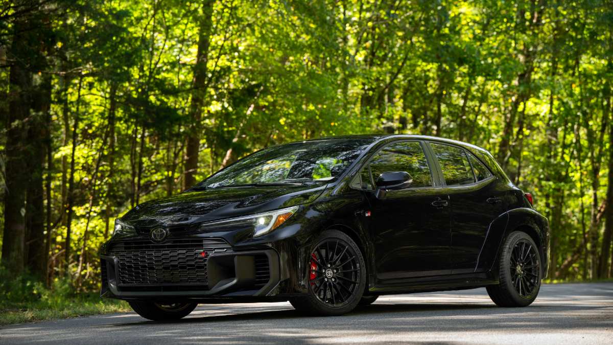 Black 2025 Toyota GR Corolla with aggressive grille, parked on road with lush green forest background, low front angle view