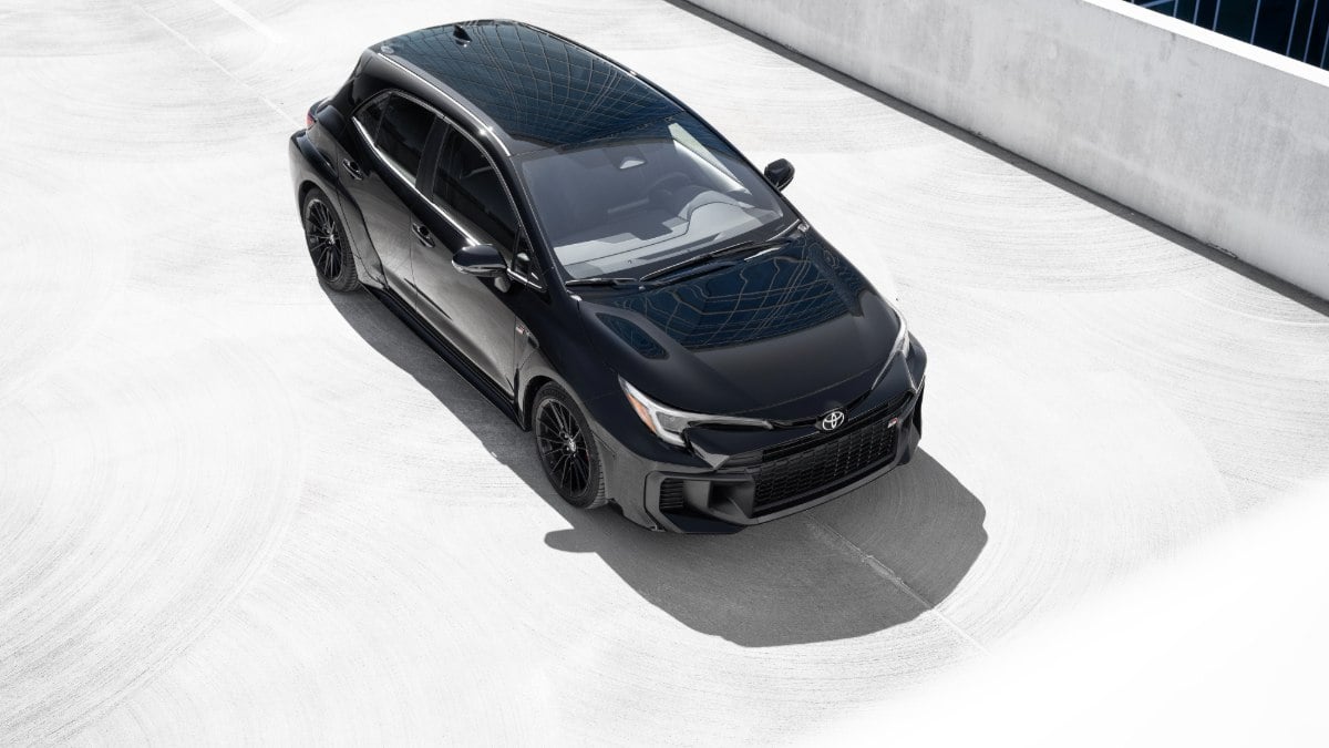 2023 Toyota GR Corolla in black, photographed from high overhead angle, featuring black alloy wheels, panoramic roof, and aggressive body styling, parked on light concrete surface.