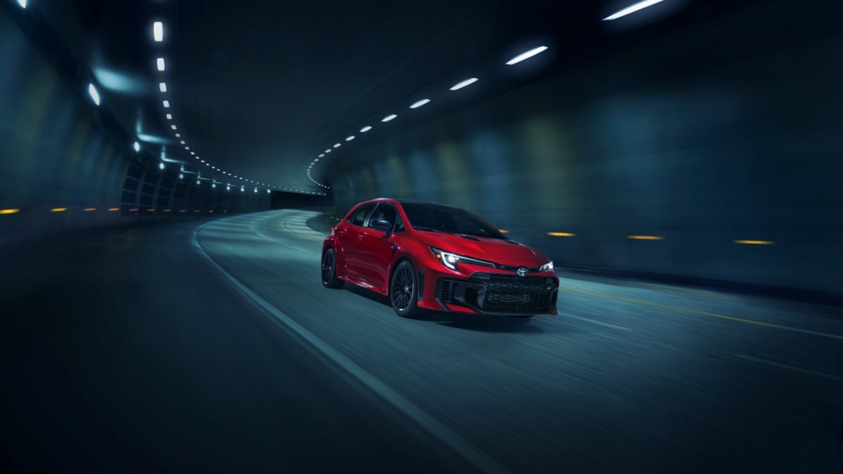2024 Toyota GR Corolla in vibrant red, photographed at three-quarter front angle inside dimly lit tunnel with dramatic overhead lighting and curved road.