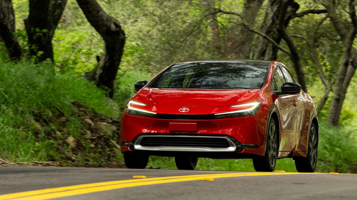 2025 Toyota Prius in red driving on a winding forest road, front three-quarter view emphasizing sporty exterior design and advanced hybrid technology.