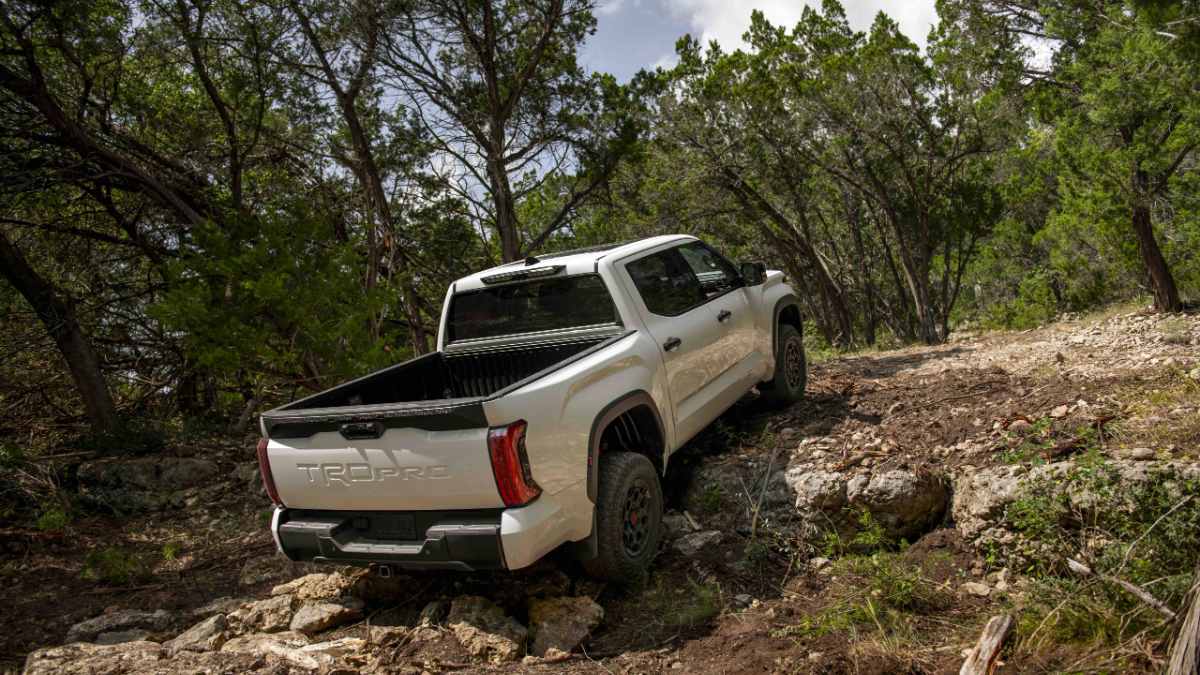 White 2024 Toyota Tacoma TRD Pro shown from rear three-quarter view, navigating rocky off-road terrain surrounded by green trees.