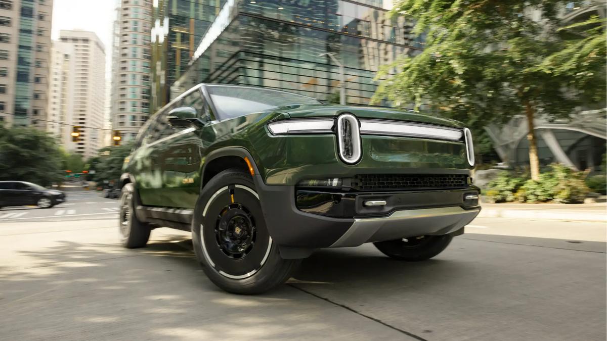 A dark green Rivian R1S electric SUV shown from the front three-quarter angle, driving on a city street with modern high-rise buildings and trees in the background.