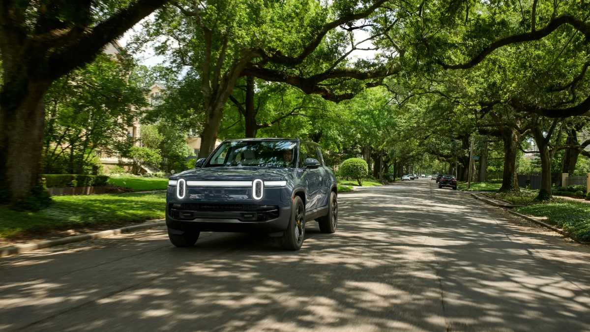 Rivian R1S electric SUV driving on a quiet, tree-lined residential street in daylight