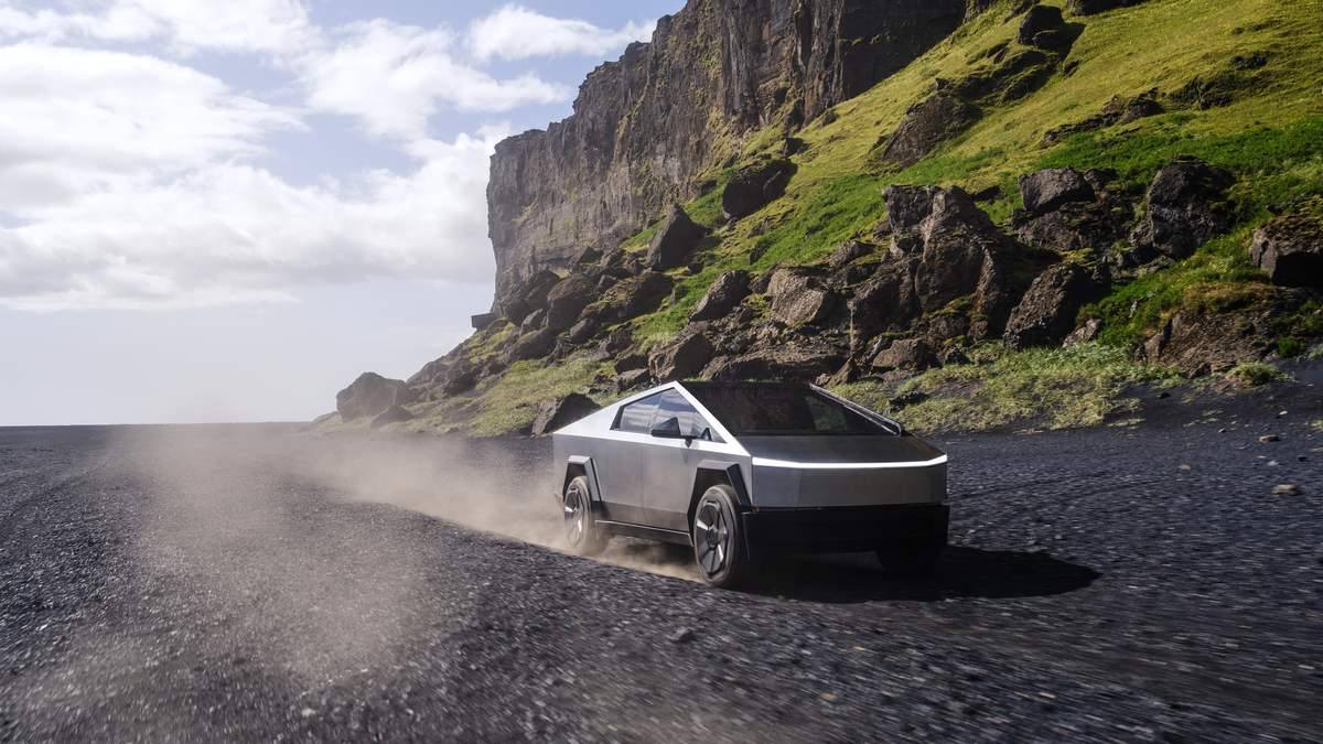 Tesla Cybertruck driving on a black sand coastal road with cliffs and rugged terrain in the background