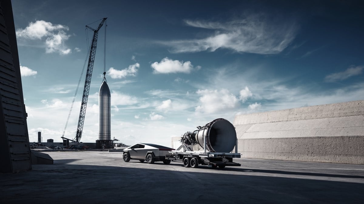 A silver Tesla Cybertruck is parked at what appears to be a SpaceX facility, with a large rocket or spacecraft component being lifted by cranes in the background against a dramatic blue sky with white clouds.