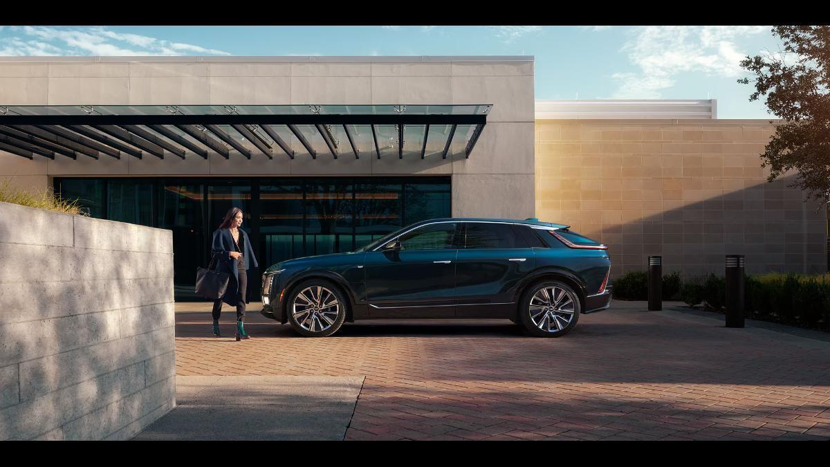 2026 Cadillac Lyriq SUV in a side view, parked with a woman walking nearby, showcasing large wheels and modern design.