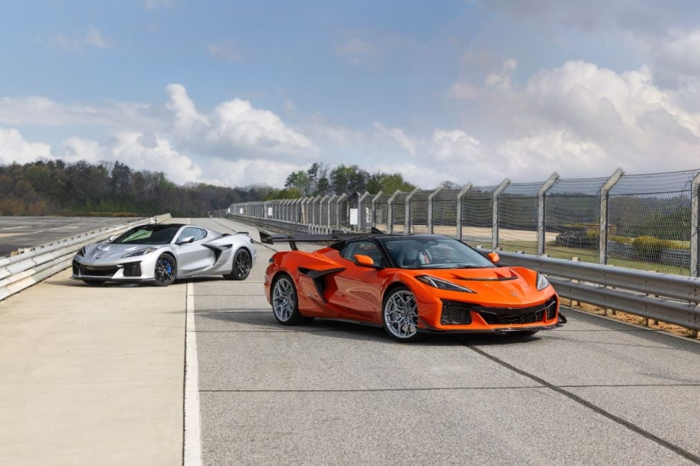 Orange and white Chevrolet Corvette Z06 convertibles, three-quarter front view, parked on racetrack with safety fencing and cloudy sky.