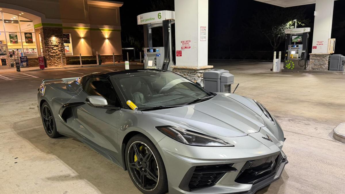 Silver Chevrolet Corvette C8 parked at a well-lit gas station at night, front three-quarter view highlighting the aerodynamic body and black wheels.