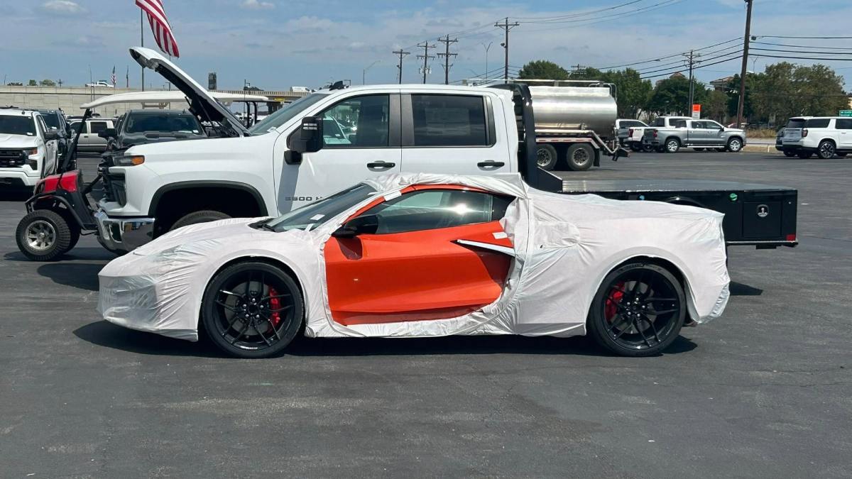 Orange sports car partially covered in protective wrap, parked beside a white truck.