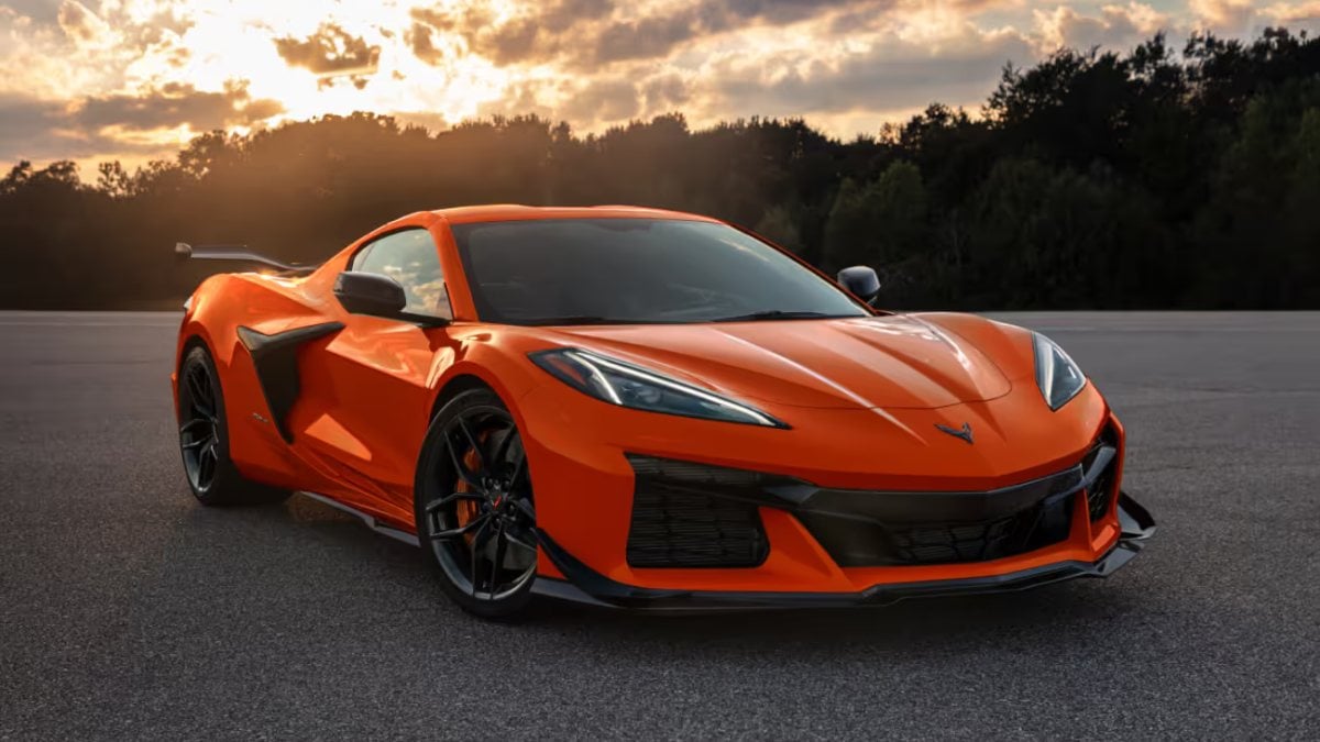 Orange Chevrolet Corvette Z06 C8, front three-quarter view, black wheels, rear spoiler, parked on asphalt at sunset with trees.