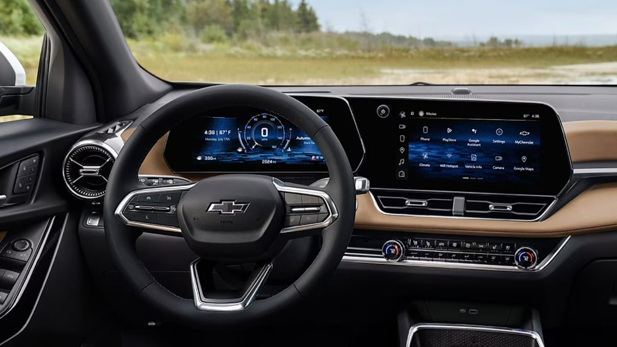 2026 Chevy Equinox EV interior with a focus on the steering wheel and two digital display screens. The screens show vehicle information and multimedia options, set against a woodsy outdoor view.