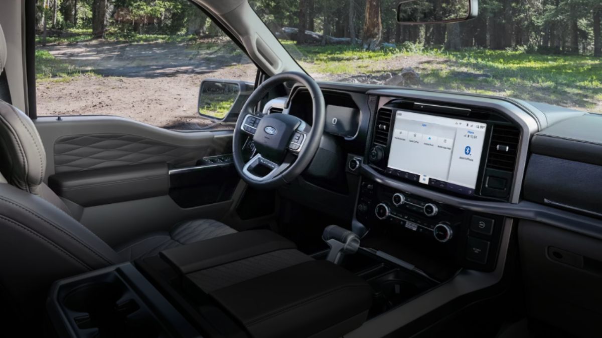 Interior view of a 2026 Ford F-150 Lightning showing the modern dashboard with large central touchscreen display, digital instrument cluster, and black leather upholstery, photographed from the passenger side with a wooded area visible through the windshield.
