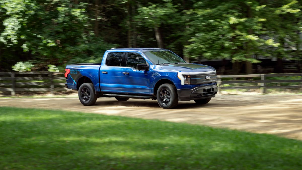 Blue Ford F-150 Lightning electric pickup truck driving on dirt road through wooded area