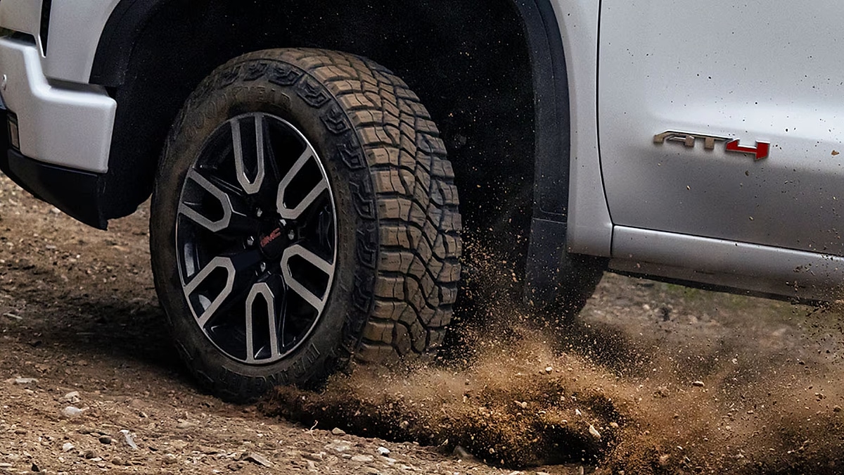 Close-up of a 2026 GMC Sierra 1500 AT4 tire on a dirt road kicking up dust. The side of the vehicle is visible with an "AT4" badge, conveying a rugged, adventurous tone.