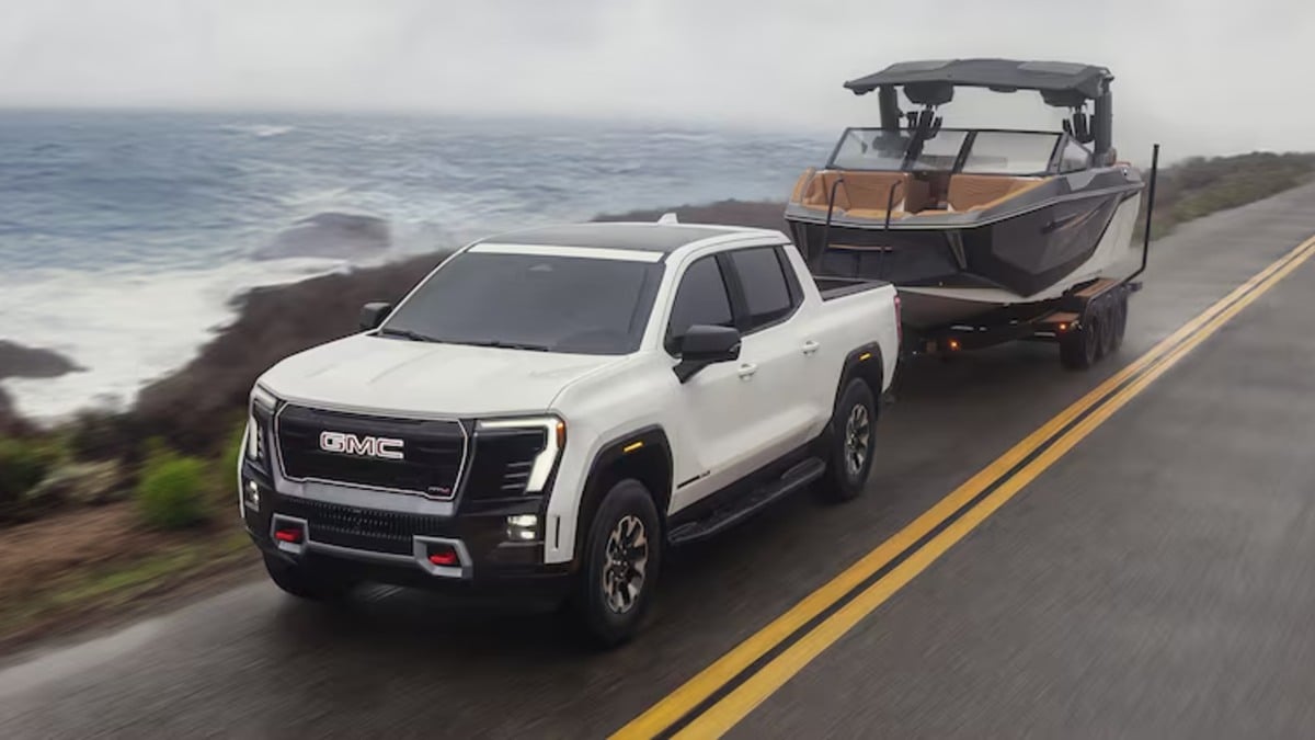 White GMC Sierra AT4X pickup truck, front three-quarter view, towing boat on coastal highway with ocean backdrop.