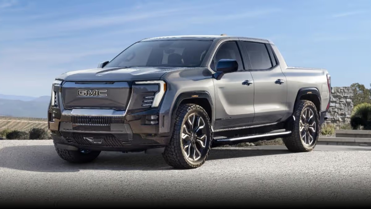 A silver 2024 GMC Sierra EV pickup truck shown from a front three-quarter angle, featuring the distinctive new front grille design and LED lighting signature against a scenic mountain backdrop.