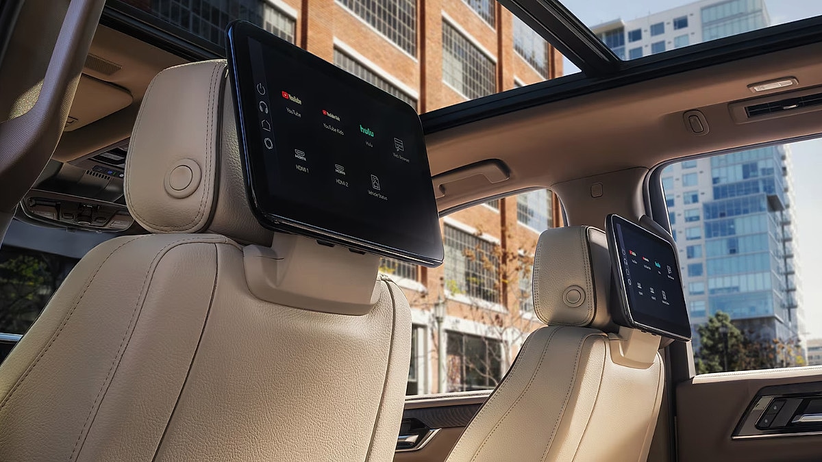 GMC Yukon Denali interior featuring beige leather seats with mounted entertainment screens displaying streaming apps. Sunroof offers a view of urban buildings.