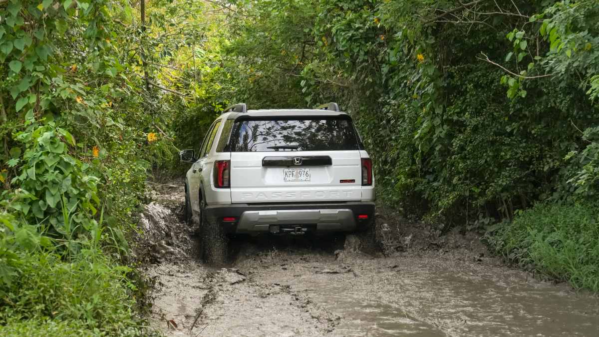 2026 Honda Passport TrailSport driving through a narrow muddy off-road trail surrounded by dense green vegetation.