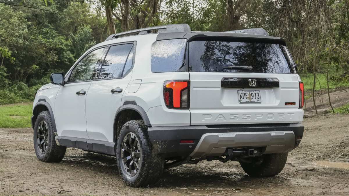2024 Honda Passport TrailSport in Platinum White Pearl, rear three-quarter view, featuring rugged all-terrain tires and raised suspension, parked on dirt trail surrounded by forest.