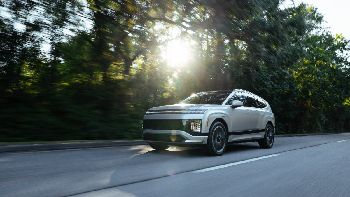 A silver Hyundai Ioniq 9 shown from a front three-quarter angle, driving on a tree-lined road with dramatic sunlight filtering through the foliage.