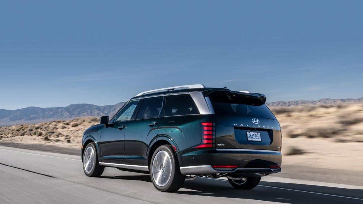 Dark green Hyundai Palisade hybrid SUV, rear three-quarter view, featuring distinctive vertical taillights, roof rails, driving on desert highway with mountains.