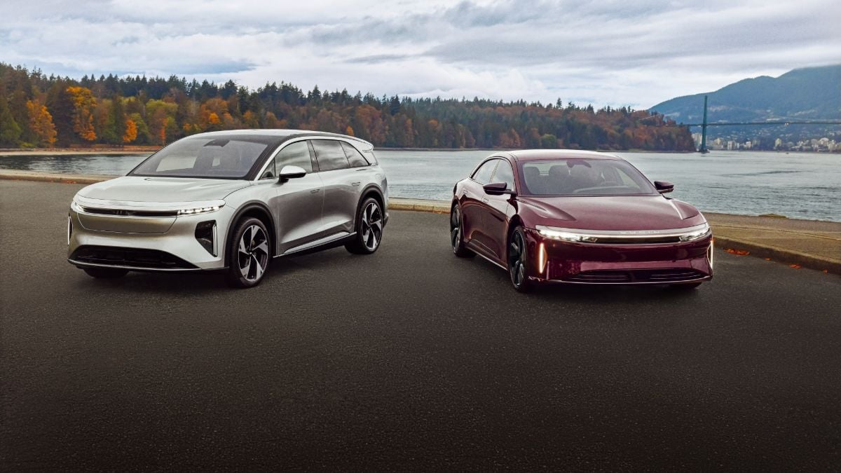 Two stylish electric vehicles, a silver SUV and a deep red sedan, parked beside a serene waterfront with colorful autumn trees in the background.