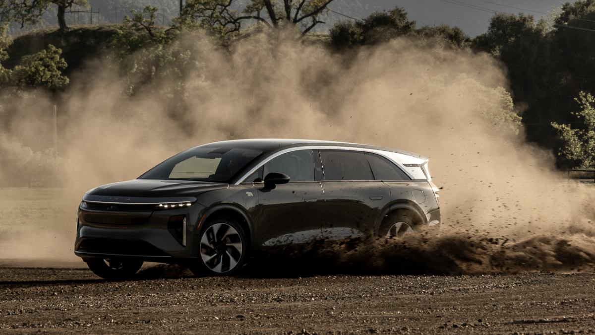 2026 Lucid Gravity electric SUV in metallic gray, shown in three-quarter view kicking up dust while off-roading on dirt terrain beneath trees.