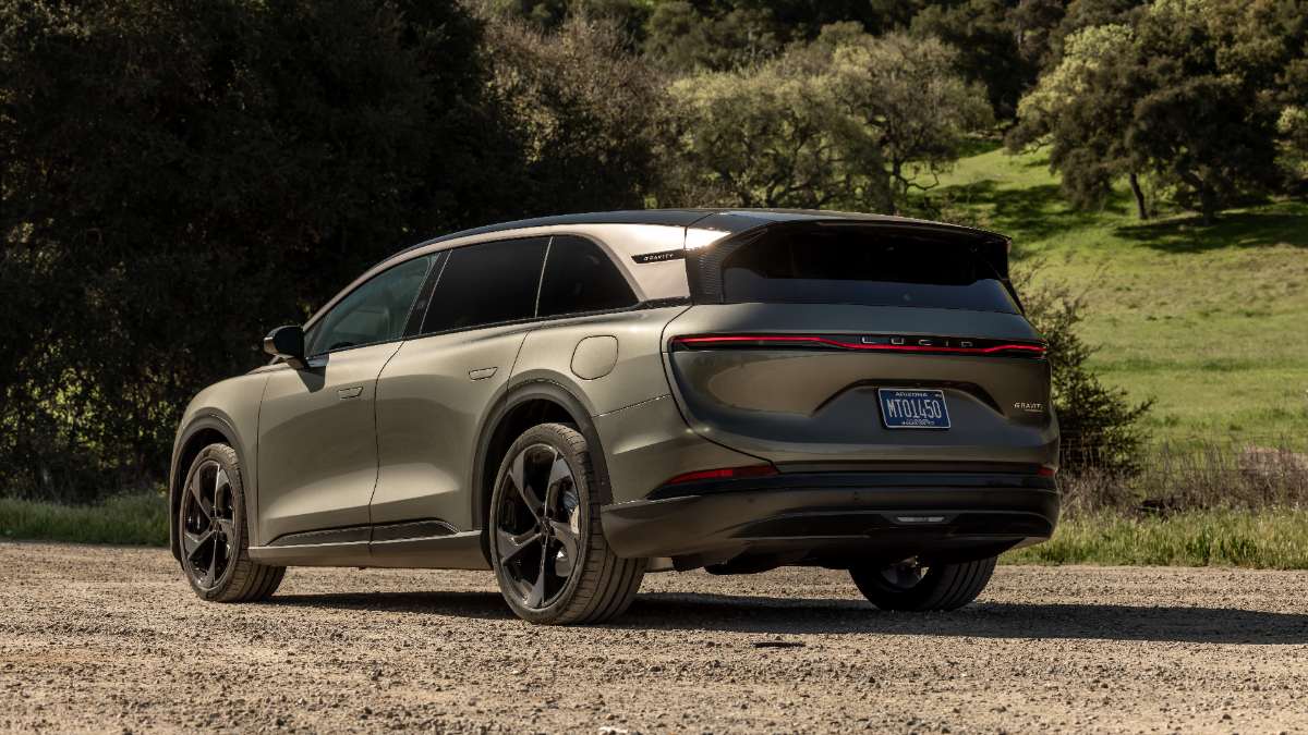 2026 Lucid Gravity SUV in matte silver, parked on gravel with green trees and grassy hills in background, rear three-quarter view