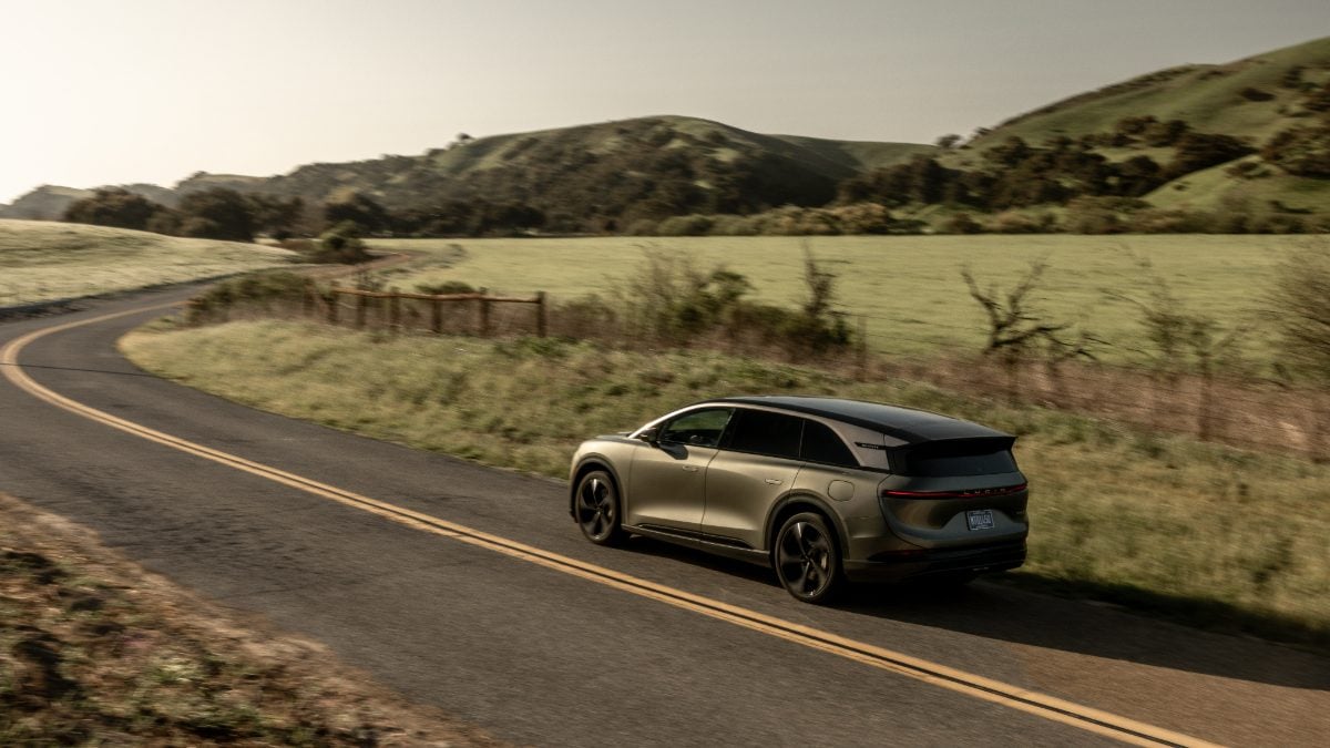 2026 Lucid Gravity electric SUV in metallic gray, shown from rear three-quarter view, driving on winding mountain road through green hillside.