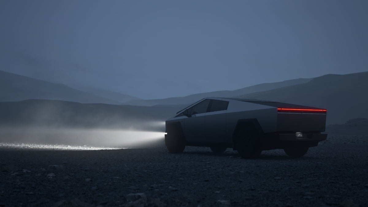 Tesla Cybertruck driving at night on a dark, rugged landscape with headlights on, showing the rear three-quarter view, stainless steel body, full-width LED taillight, and off-road capability in low-light conditions.