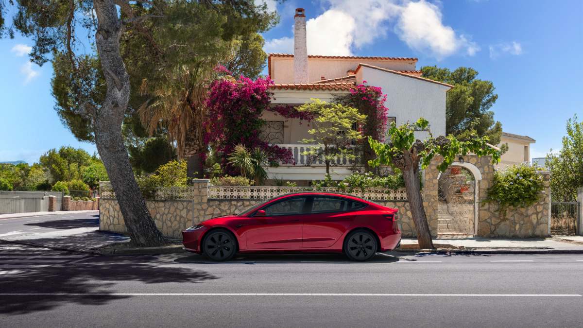A red Tesla Model 3 is parked on a street side view, with a Mediterranean-style house featuring bougainvillea flowers and stone walls in the background under bright blue skies.
