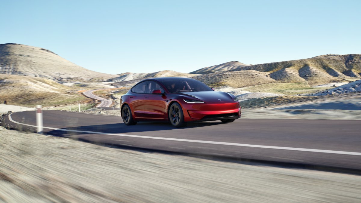 Red electric car driving on a winding road through a hilly, desert landscape.