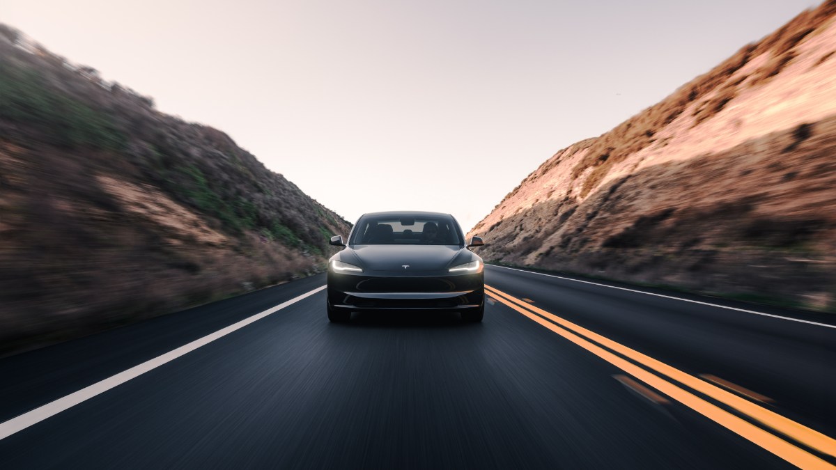 Black Tesla Model 3, front view, driving on winding mountain road with illuminated LED headlights against dramatic rocky cliffs.