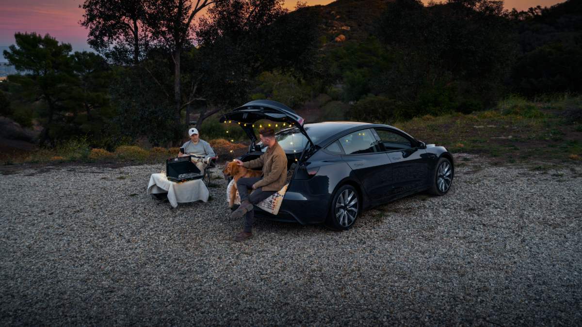 Black Tesla Model 3 parked at dusk campsite with string lights, people relaxing by trunk, dog present, gravel ground, wooded setting.